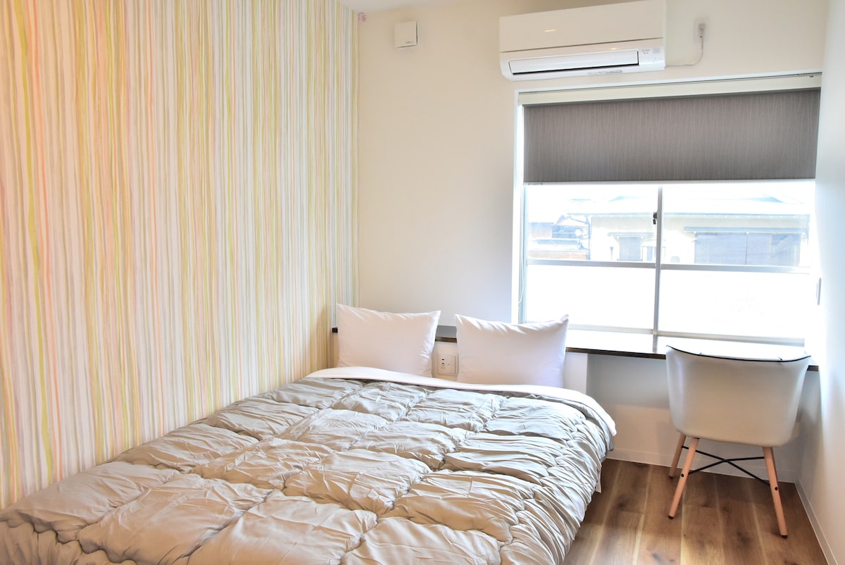 A cozy sleeping area is presented with a spacious bed covered by a soft comforter, complemented by two pillows. A window allows natural light to fill the room, and a simple chair is positioned at a small desk, enhancing functionality.