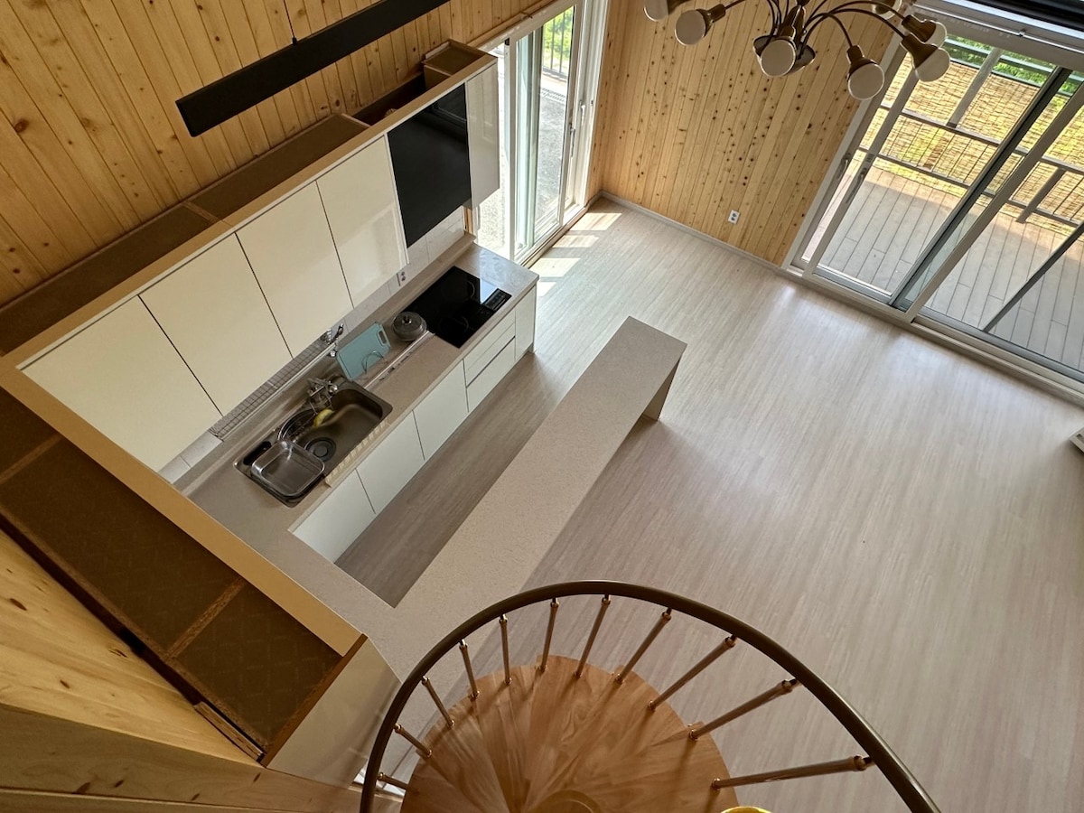 A modern kitchen space features sleek white cabinetry and a countertop extending into a spacious open area. Large glass doors allow natural light to enter, while a spiral staircase provides access to the upper level. Natural wood paneling adds warmth to the environment.