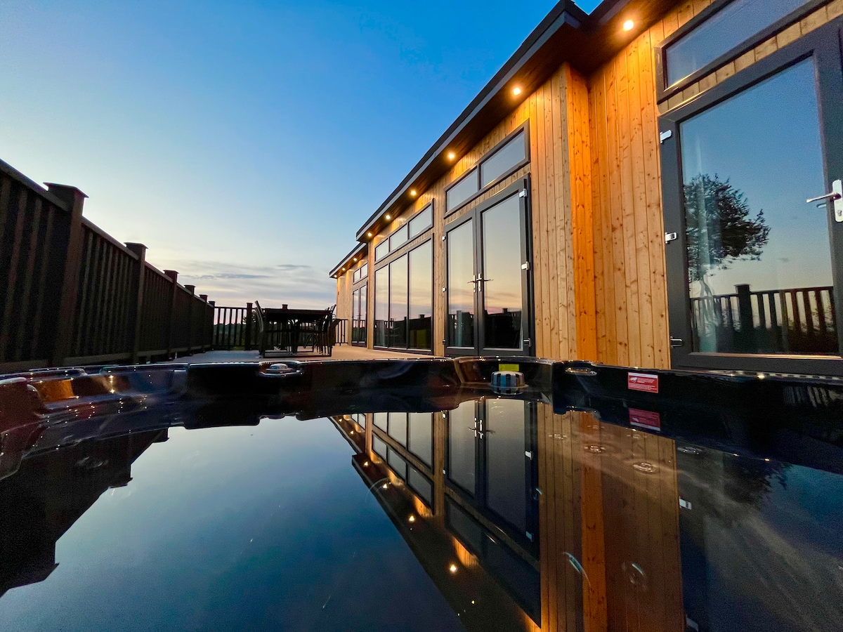 The exterior of the lodge is shown, with large glass doors reflecting the evening sky. The wooden structure is warmly illuminated, and a hot tub is visible in the foreground, mirroring the surroundings. The railing hints at the garden area beyond.
