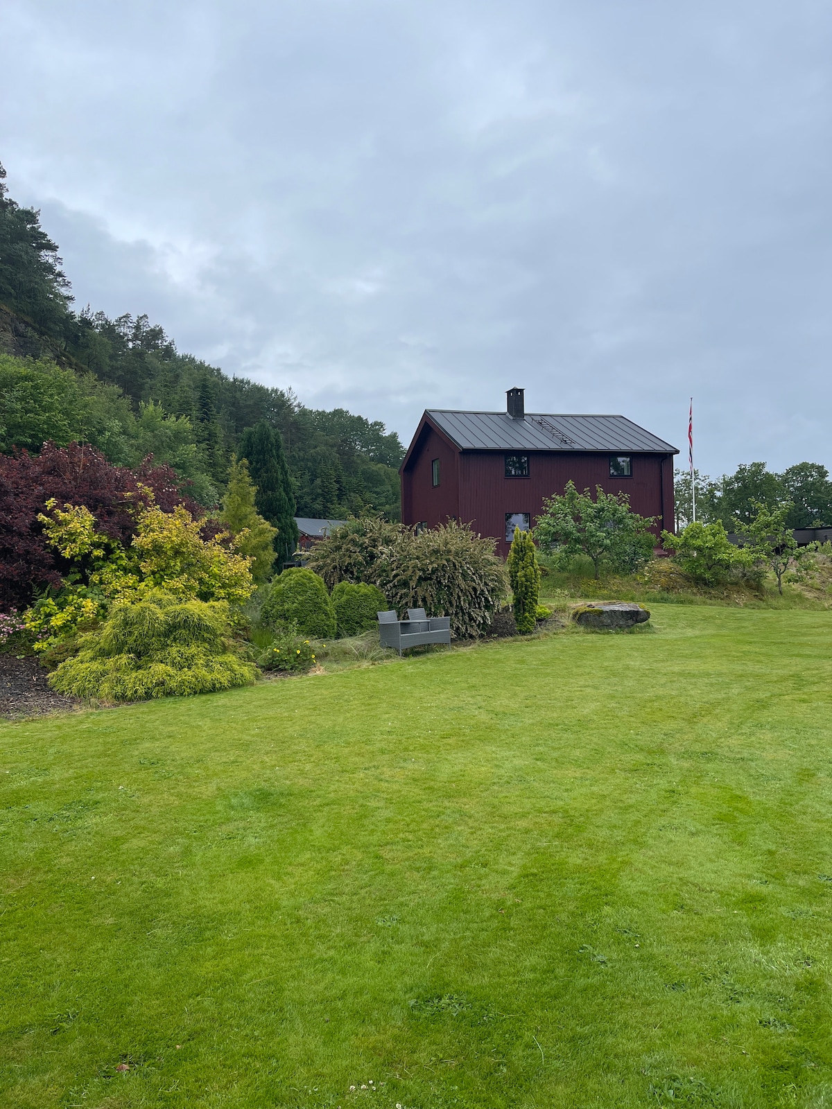 A charming red house is nestled among lush greenery and vibrant bushes. The expansive lawn is neatly maintained, with varied plant life providing color and texture. A seating area is positioned in the garden, inviting relaxation amidst the serene natural surroundings.