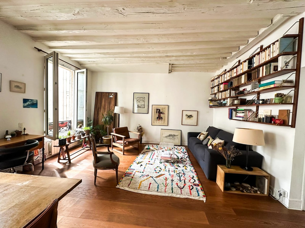 The living area is illuminated by natural light streaming through large open windows. A dark sofa faces a decorative wall shelf filled with books, while colorful artwork adorns the walls. A plush, patterned rug lies on the wooden floor, complementing the cozy seating arrangement.