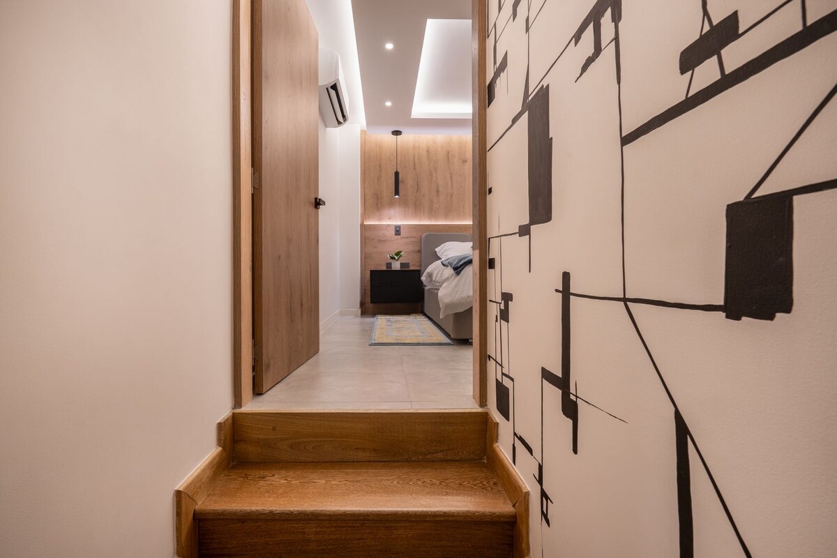 A staircase leads to an upper-level area, with a modern design featuring a softly lit pathway. The walls are adorned with abstract black artwork, providing a contemporary backdrop to the airy space with a cozy bedroom visible at the top.