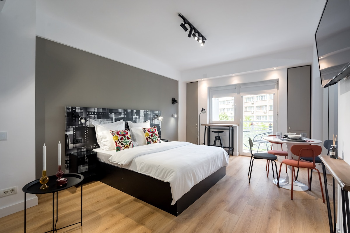 The studio features a spacious bed with decorative cushions against an artistic black and white headboard. A dining area with colorful chairs is positioned nearby, with a small table and a scenic balcony view through large glass doors. Natural light fills the room.