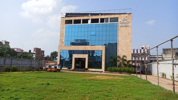 A modern building with a reflective glass facade, showcasing a blend of stone and metal materials. A spacious green lawn in front offers an open area, with a view of surrounding structures in the distance. Clear skies accentuate the building's contemporary design.