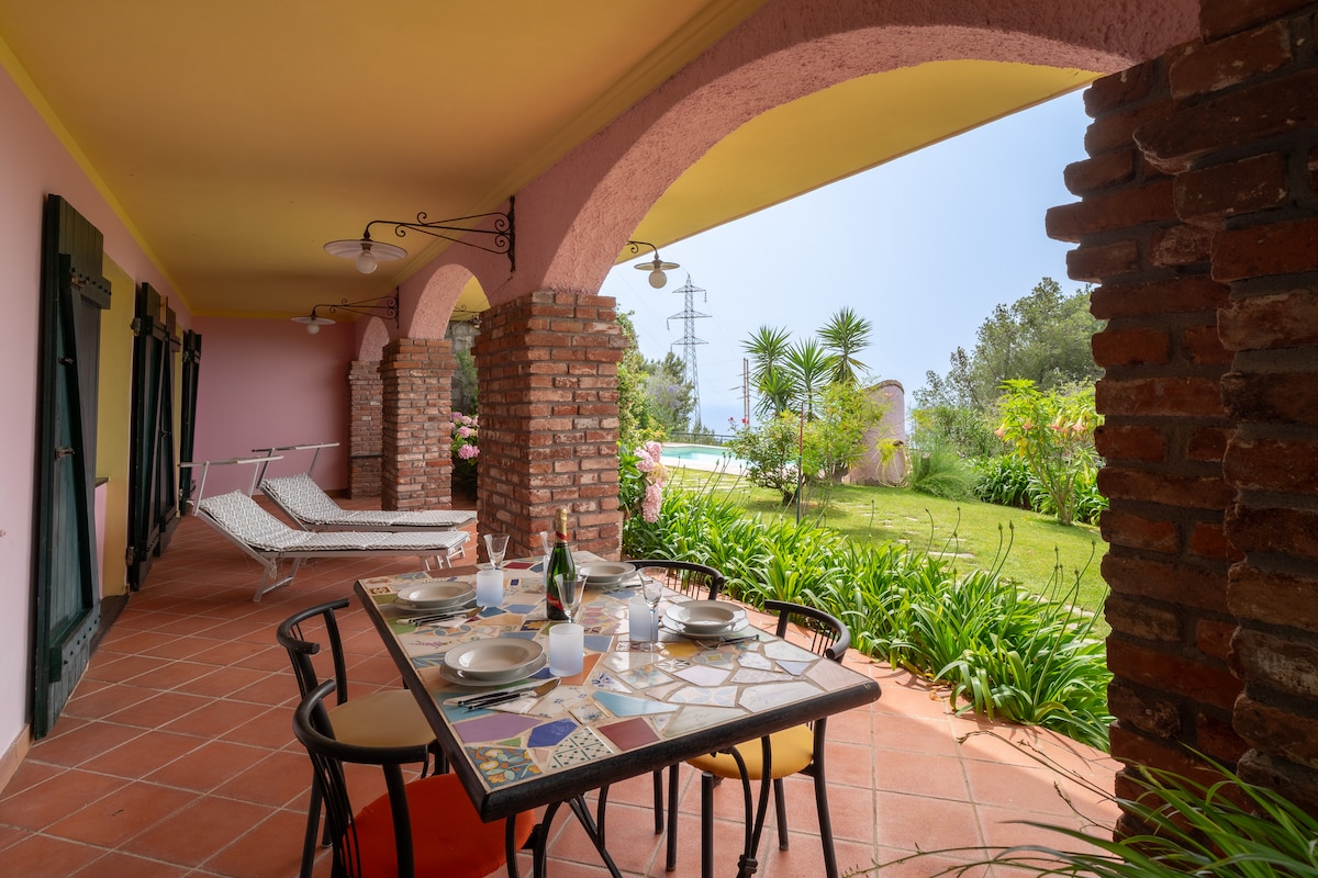 A covered terrace features a dining table set with plates and glasses, surrounded by lush greenery and planters. Two loungers are positioned nearby, ideal for relaxation. In the background, a swimming pool is visible, framed by well-maintained grass and colorful plants.