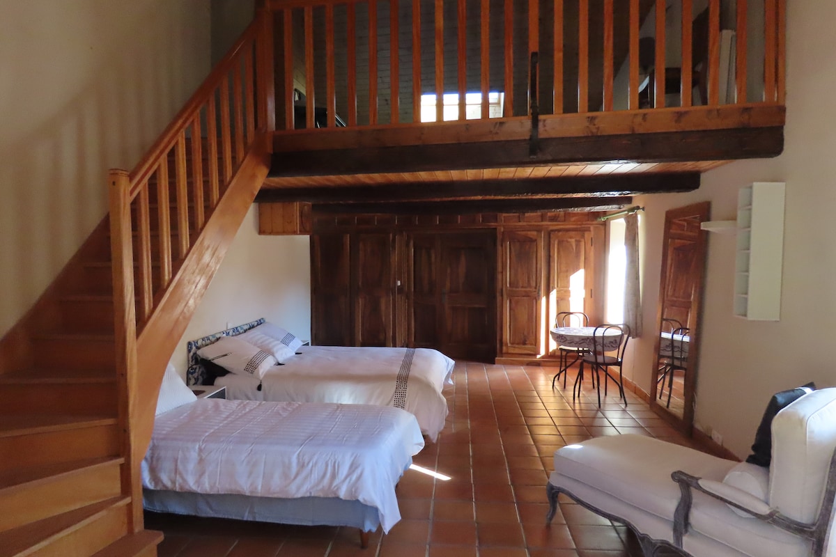 A spacious mezzanine room features wooden beams and a warm tiled floor. Two beds are positioned on one side, with a table and chairs nearby. A staircase leads to the upper level, while large windows provide ample light and views of the surrounding area.
