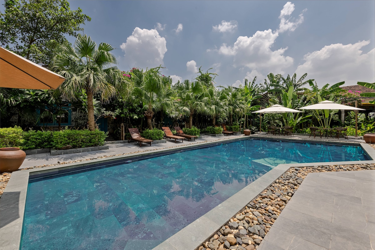 A spacious outdoor pool is surrounded by lush greenery, with palm trees and vibrant plants visible in the background. Lounge chairs are positioned on one side, and shaded umbrellas offer respite from the sun, creating an inviting outdoor relaxation area.