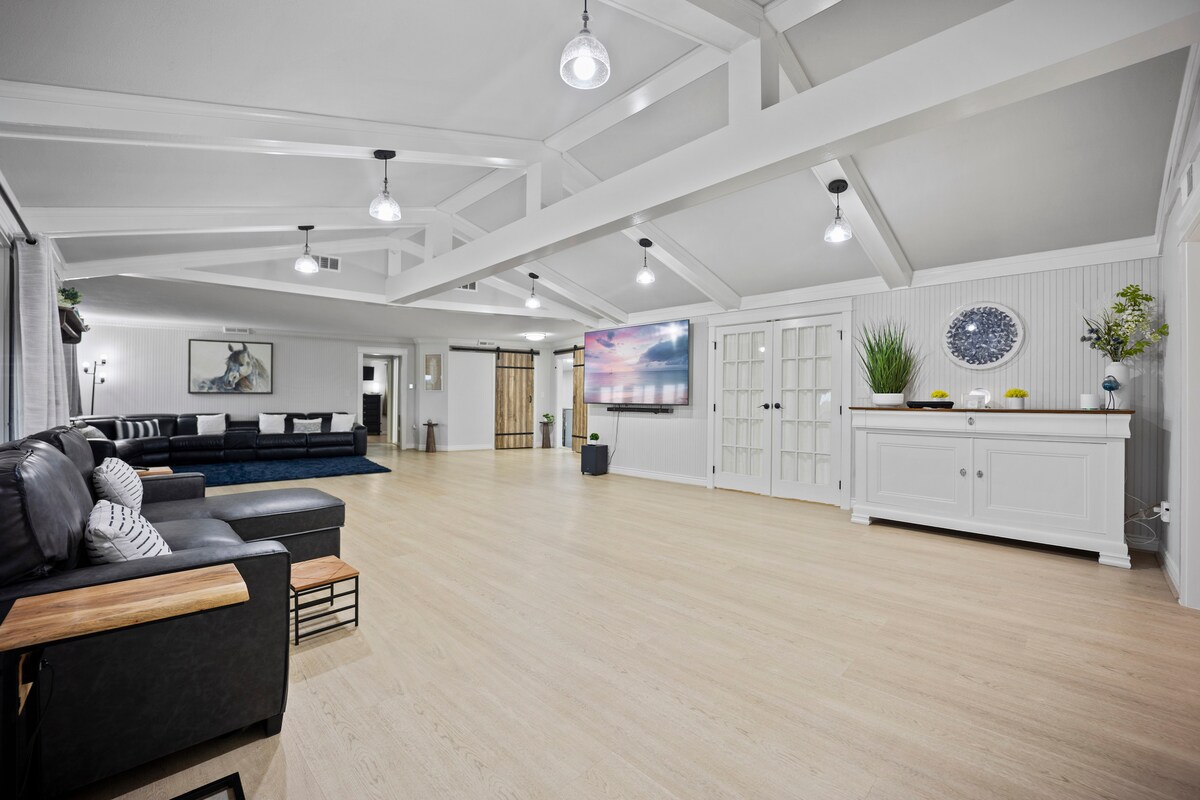 A spacious living room is presented, featuring high vaulted ceilings supported by exposed beams. A large sofa is positioned in one corner, complemented by a wooden table. An oversized smart TV is mounted on the wall, providing an ideal area for relaxation and entertainment.