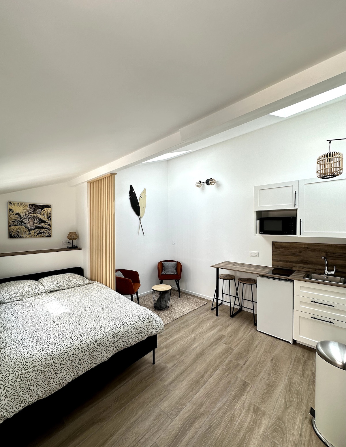A studio space features a spacious double bed with a patterned duvet. Two modern red chairs are paired with a small side table, while a compact kitchenette displays a microwave and stove. Light pours in from above, enhancing the open layout with neutral tones.