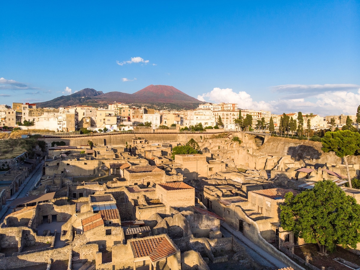 The image captures an aerial view of historical ruins nestled against a backdrop of Mount Vesuvius. Stone structures, some with tiled roofs, are interspersed among trees, while a nearby town features low-rise buildings under a clear blue sky, enhancing the landscape's rich history.