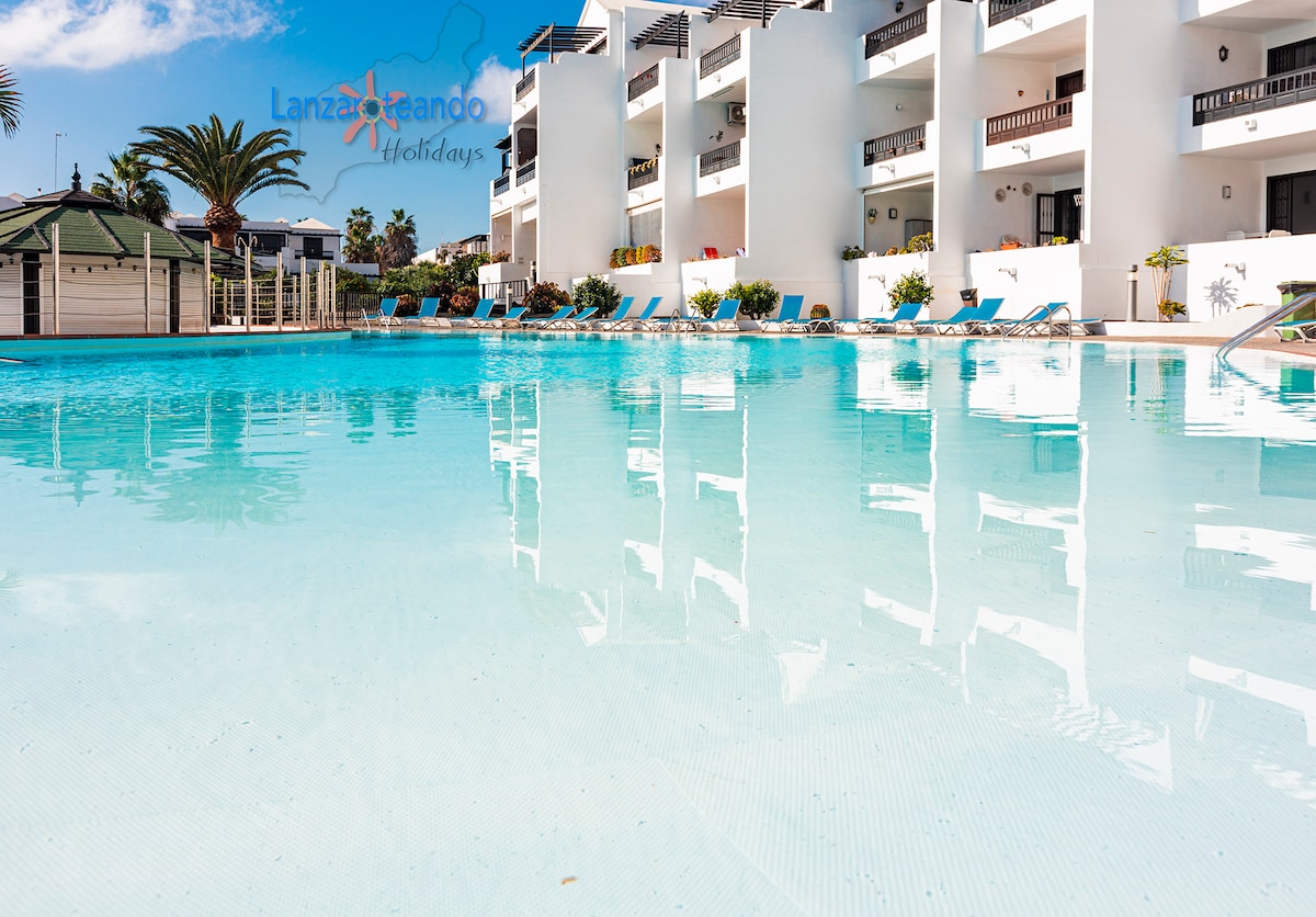 A tranquil communal pool area is depicted, with clear blue water reflecting the surrounding palm trees. Lounge chairs are arranged neatly along the poolside, providing a relaxing atmosphere. The white building with balconies is visible in the background, complementing the well-kept landscape.