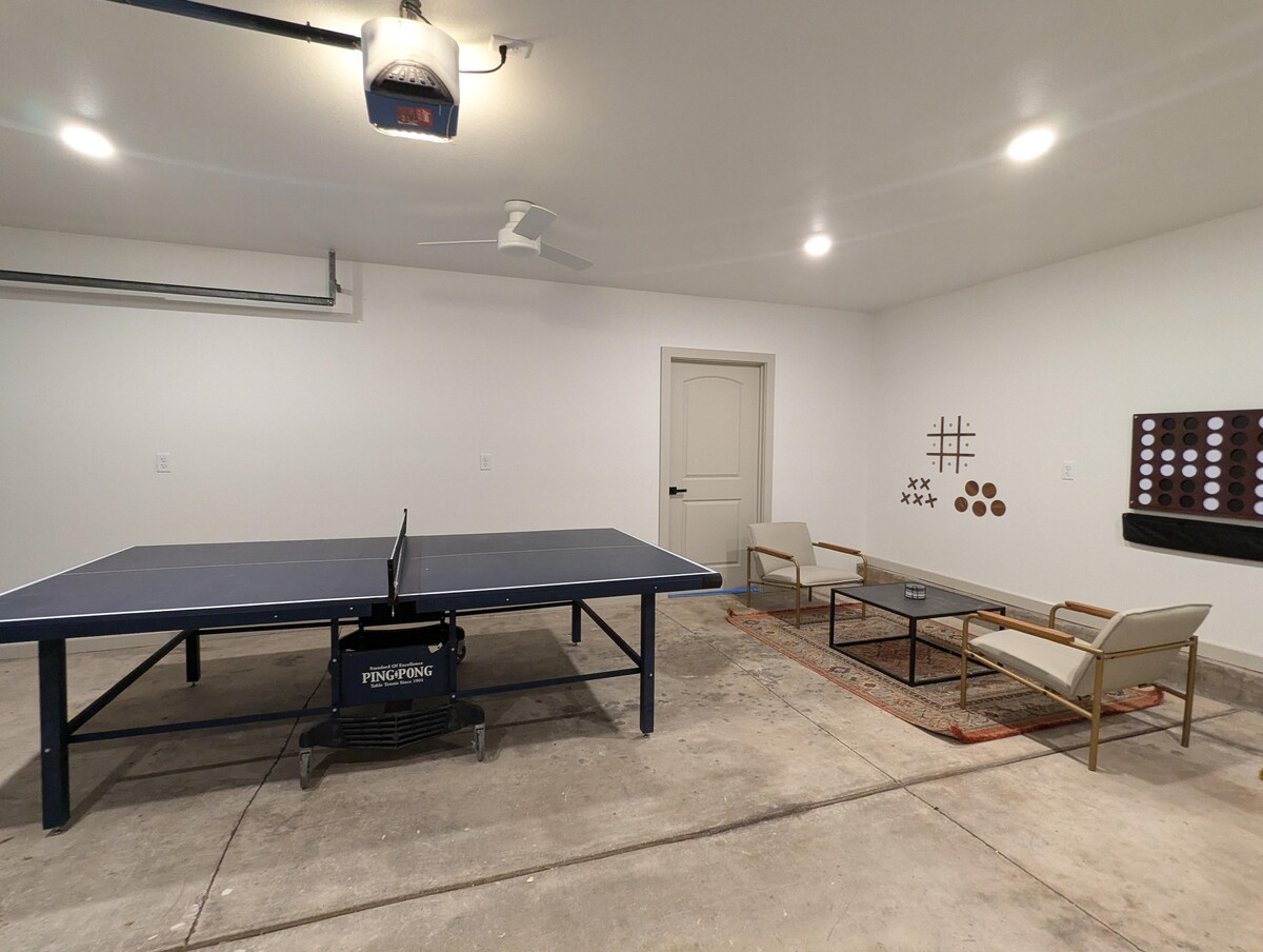 A spacious garage converted into a game room includes a table tennis table and a seating area with two chairs and a coffee table. The walls are decorated with a simple pattern, and a small rug is placed beneath the furniture. Ample lighting and a ceiling fan enhance the space.
