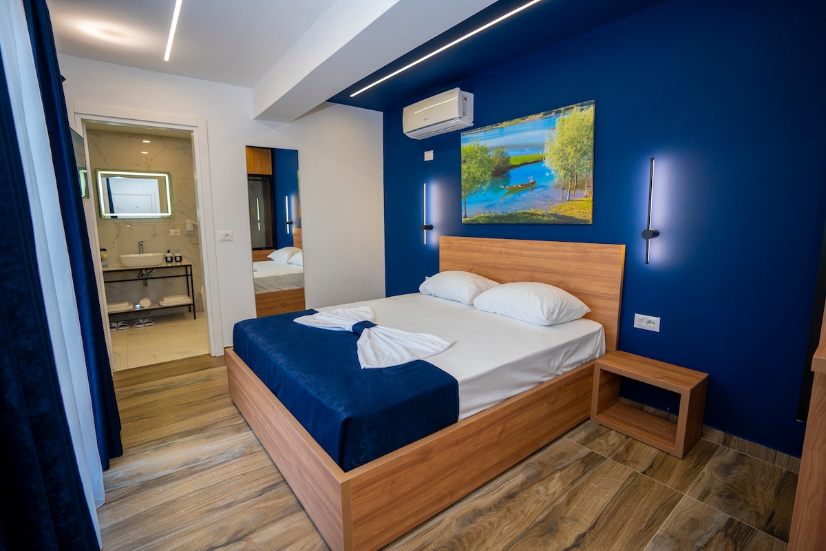 A cozy bedroom features a spacious bed with white linens, complemented by a dark blue throw blanket. The room is painted in a deep blue hue, and a landscape artwork adorns the wall above the bed. A wooden nightstand sits beside the bed, and an ensuite bathroom is visible through an open door.