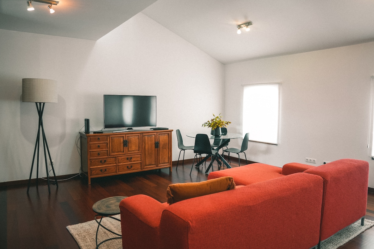 A spacious living area is furnished with a bold red sofa facing a television. A retro wooden cabinet stands against a wall, while a circular dining table with chairs occupies one corner. Natural light enters through a window, illuminating the polished wooden floor.