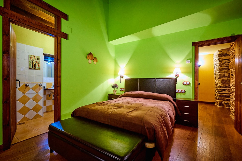 A bedroom is featured with green walls and wooden accents. A large bed is positioned against the wall, complemented by nightstands with lamps. The wooden floor adds warmth, while a bench at the foot of the bed provides additional seating. A doorway leads to a bathroom area.
