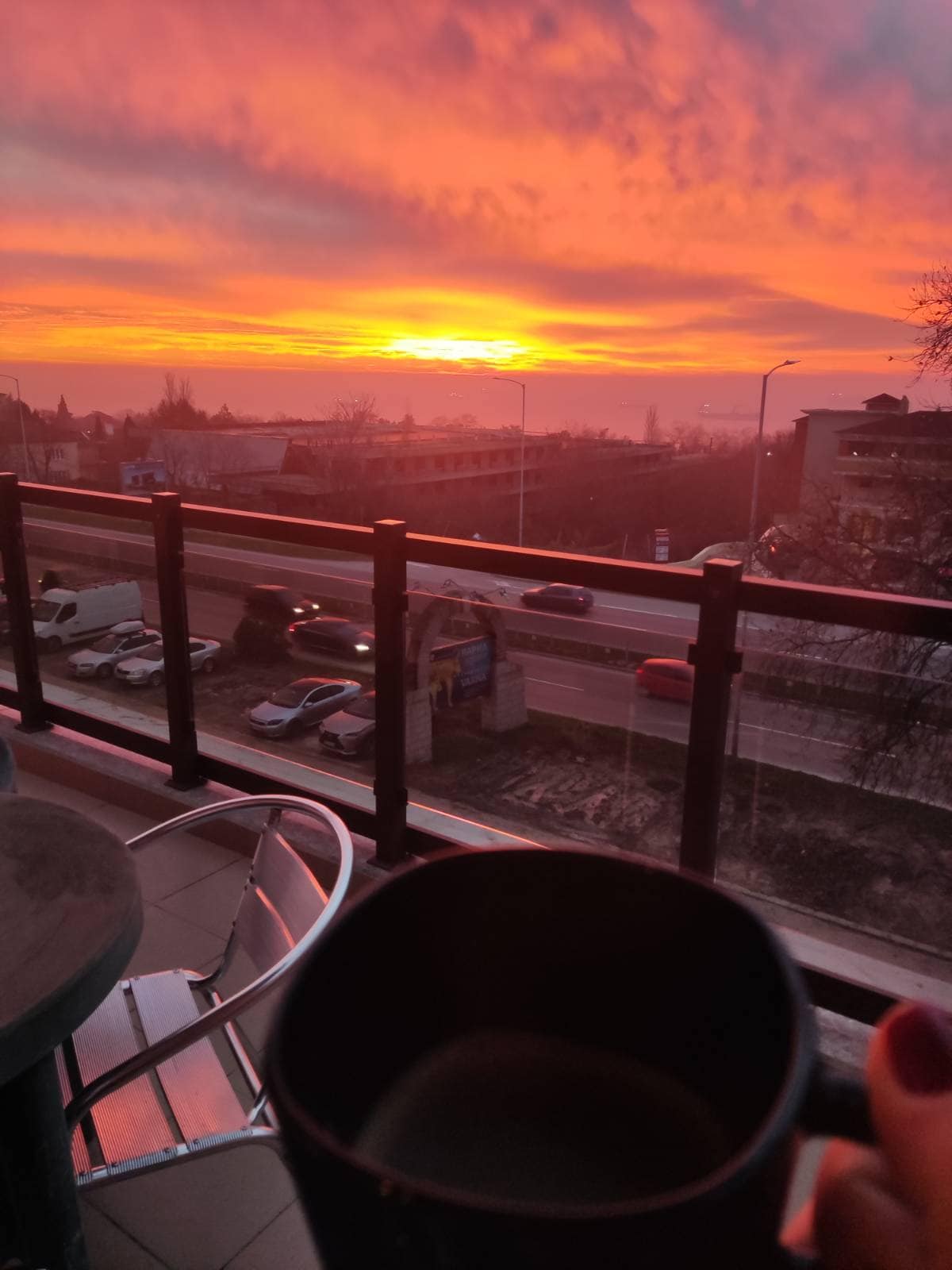 A stunning sunset fills the sky with vibrant orange and pink hues, viewed from a balcony. A sleek railing frames the scene, while distant cars move along a road below. A hand holding a dark mug is visible, suggesting a moment of relaxation.