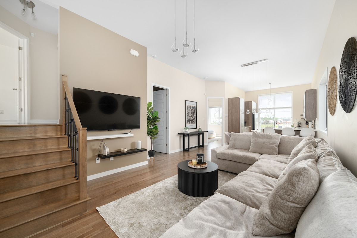 A spacious living area features a large sectional sofa and a round coffee table, complemented by a flat-screen television mounted on the wall. Natural light floods the space through multiple windows, highlighting the warm neutral tones of the walls and furnishings. A staircase is visible in the background.