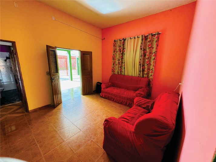 The living area features vibrant orange and pink walls, with two red sofas positioned across from one another. Light floods in through an open door, highlighting the tiled floor and colorful curtains that frame the windows.