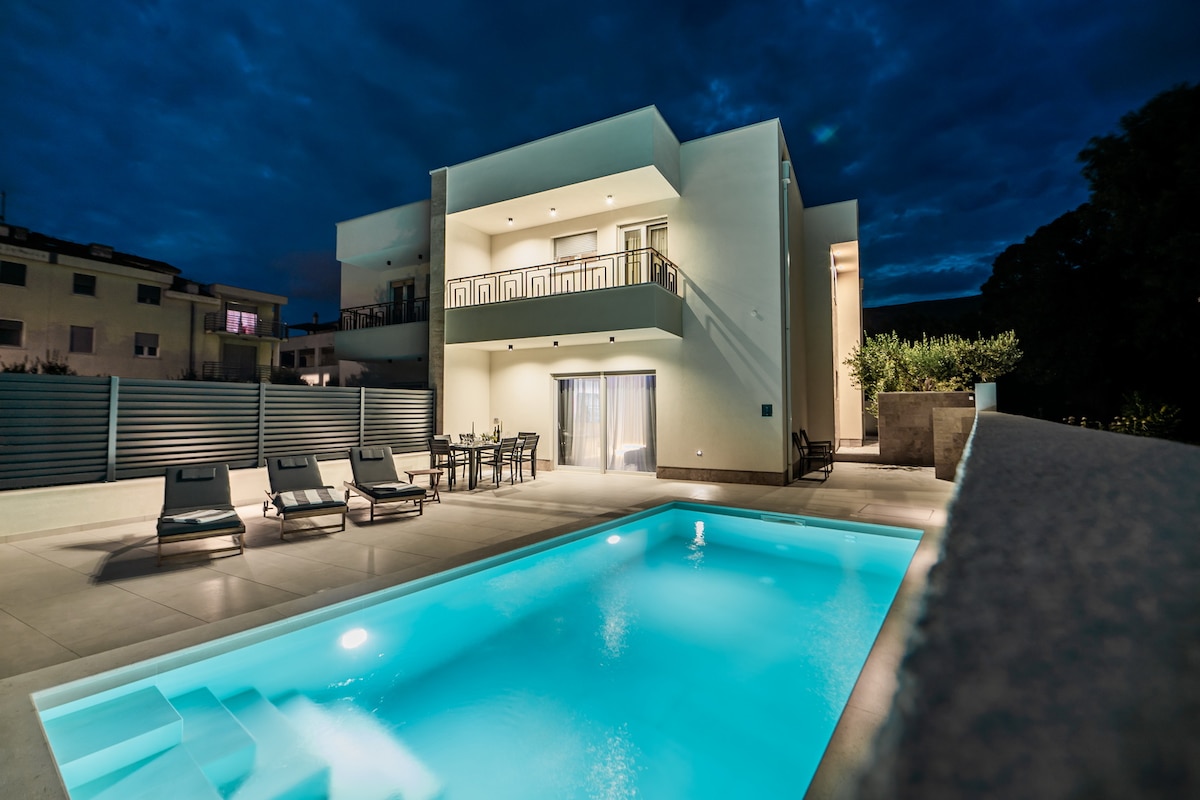 A modern villa is showcased at night, featuring a well-lit outdoor pool with soft turquoise water. Comfortable lounge chairs are arranged around the pool area, while a dining set is visible on the patio. The building has multiple levels and large windows that reflect the evening sky.
