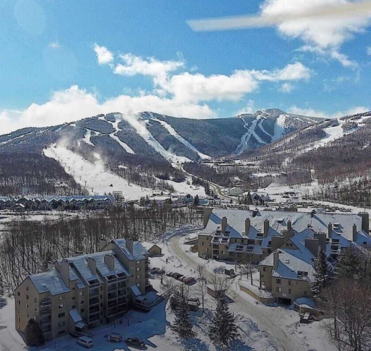 Aerial view of the Mountain Green community, surrounded by snow-covered slopes. The architectural layout is visible, with buildings situated along a winding road. In the background, ski trails are defined by their white cover, contrasting against the blue sky dotted with clouds.