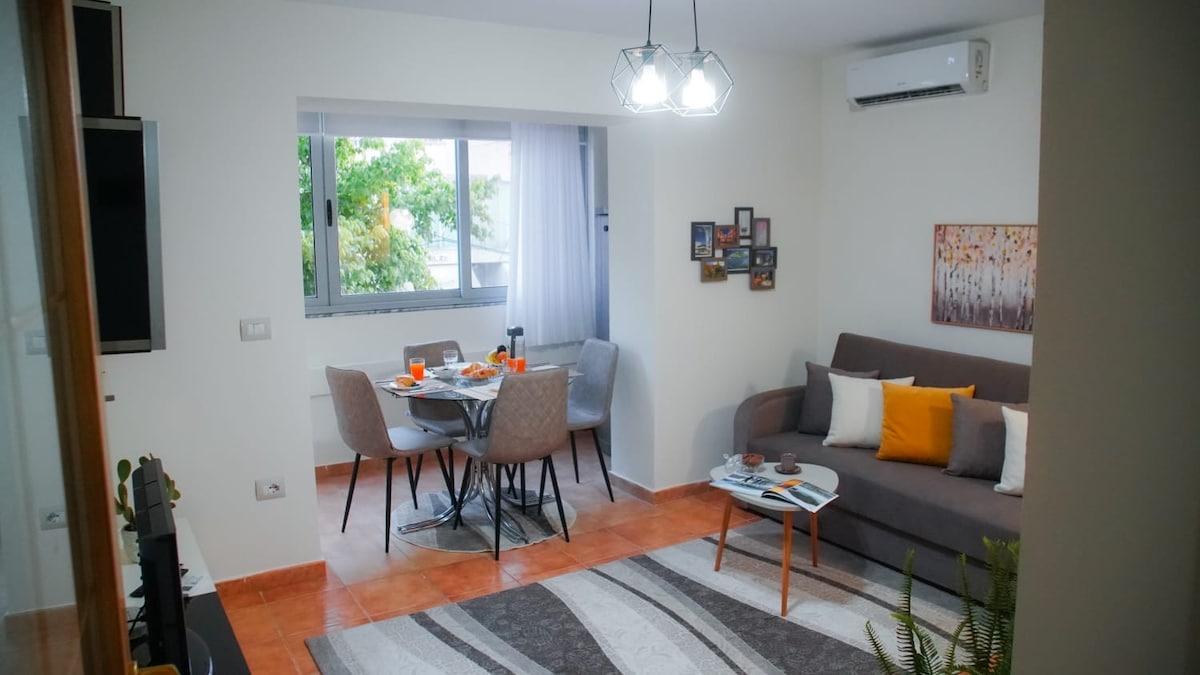 A modern living and dining area is visible, featuring a spacious sofa with throw pillows, a coffee table, and an air conditioning unit. A dining table with four chairs is positioned by a large window providing natural light, complemented by decorative wall art.