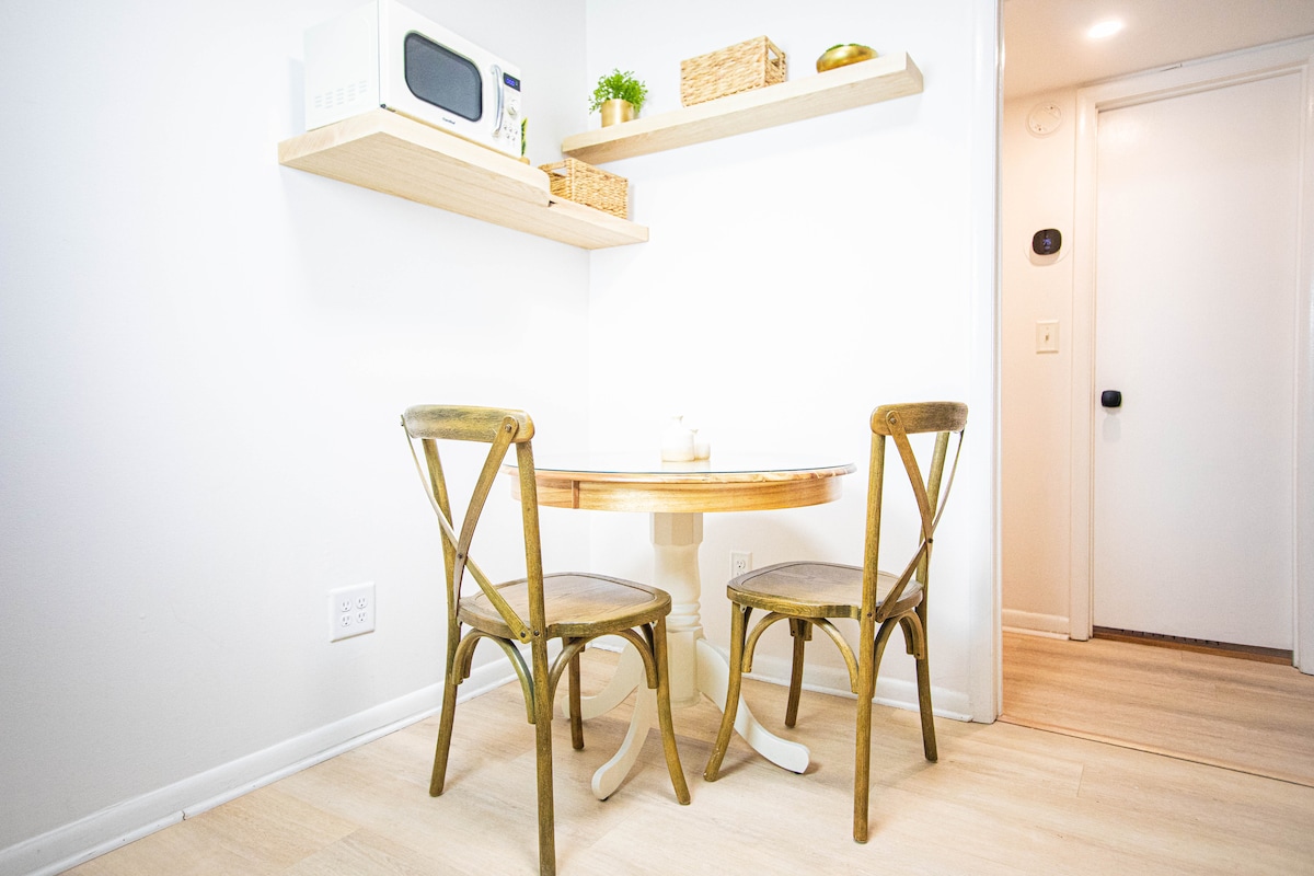 A compact dining area features a round table accompanied by two wooden chairs. A small shelf displays a microwave and decorative items, while light-colored walls and flooring enhance the bright atmosphere. The entryway is visible in the background, offering easy access.