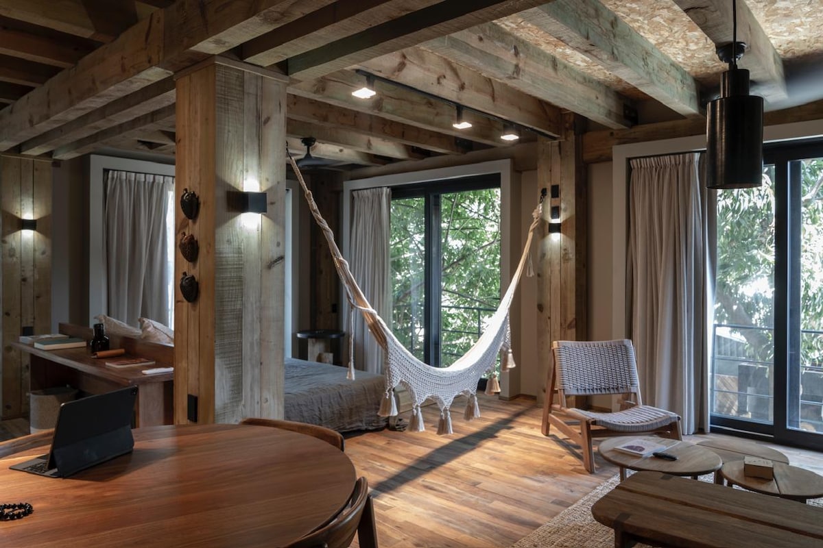 The studio showcases an open layout with exposed wooden beams and rich wood flooring. A traditional hammock is suspended near large glass doors that lead to outside greenery, while a comfortable woven chair and desk are positioned beside the spacious bed, enhancing the inviting atmosphere.