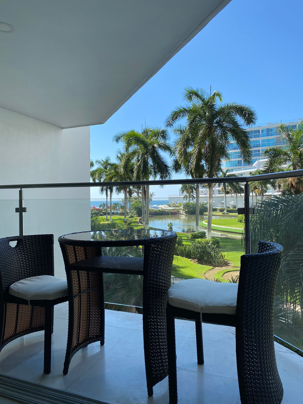 A private balcony is depicted with two high-backed wicker chairs and a small round table. Lush palm trees frame the view, revealing a glimpse of the well-maintained gardens and the distant ocean under a clear blue sky.
