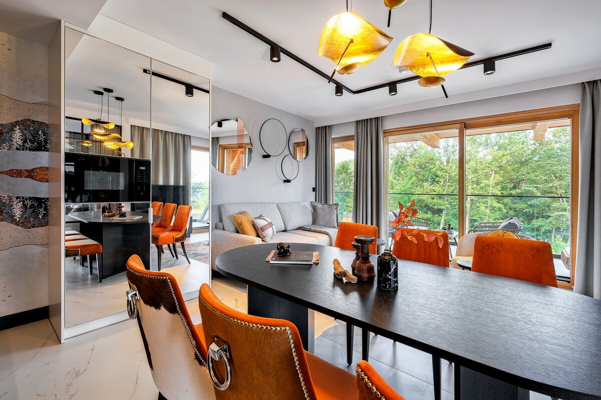 A modern dining area features a sleek black table surrounded by stylish orange chairs. A large mirror reflects a bright living space with comfortable seating. Natural light pours in through expansive windows, offering views of greenery outside. Unique light fixtures add a contemporary touch.