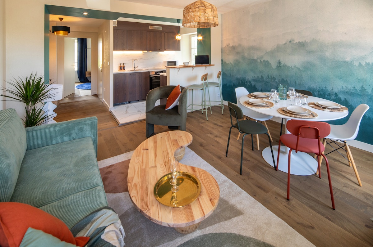 A spacious living area features a comfortable green sofa alongside a unique wooden coffee table. A dining table is set with elegant tableware, surrounded by colorful chairs. Behind the dining area, an open kitchen and a softly painted mural create a harmonious ambiance.