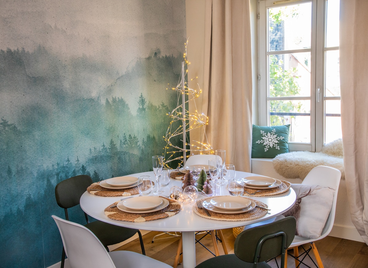A dining area is highlighted by a round table, set for six with dishes and glassware. A soft green landscape mural serves as a backdrop, while fairy lights adorn a small tree. Natural light enters through the window, enhancing the cozy atmosphere.