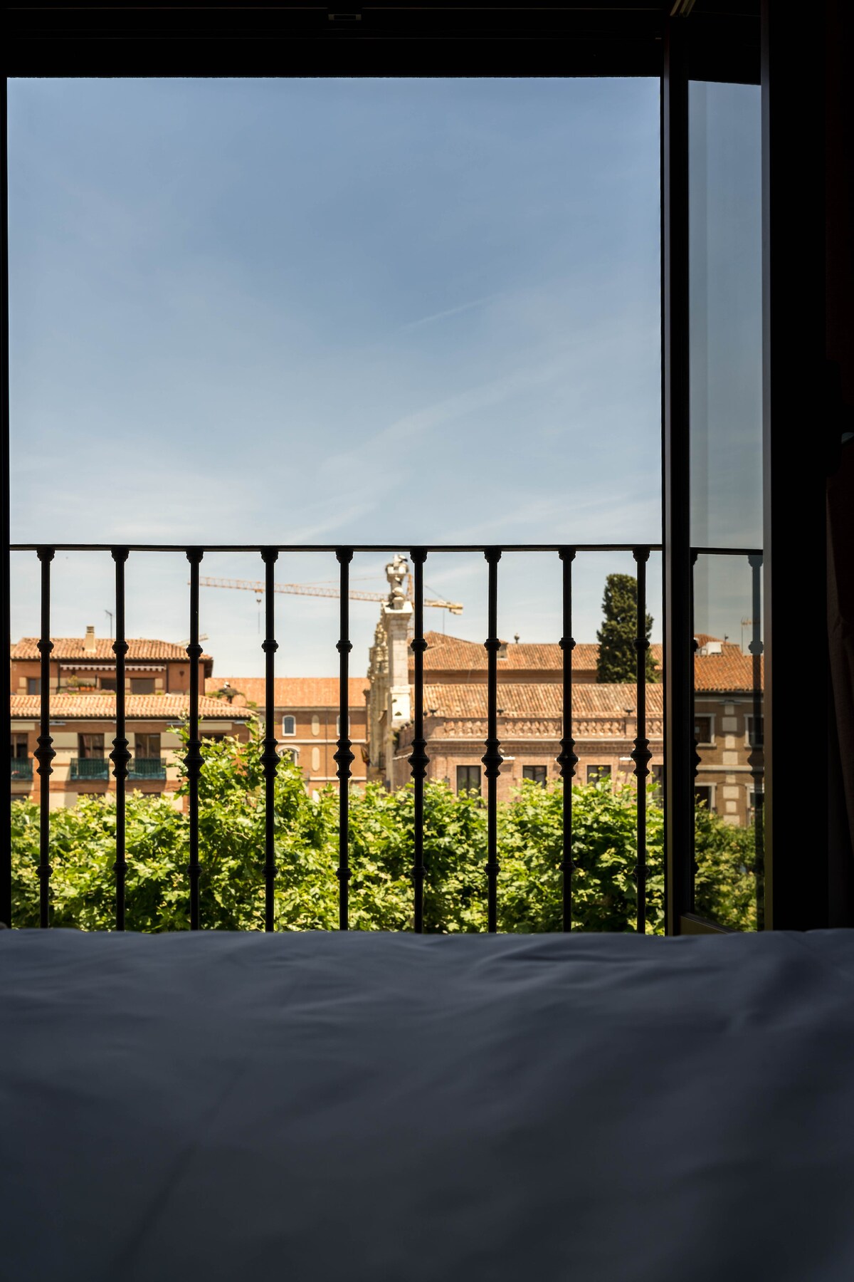 A view through the balcony reveals the vibrant greenery below and a glimpse of Plaza Cervantes. The iconic statue of Miguel de Cervantes stands prominently against a clear blue sky, framed by the building's elegant railing.