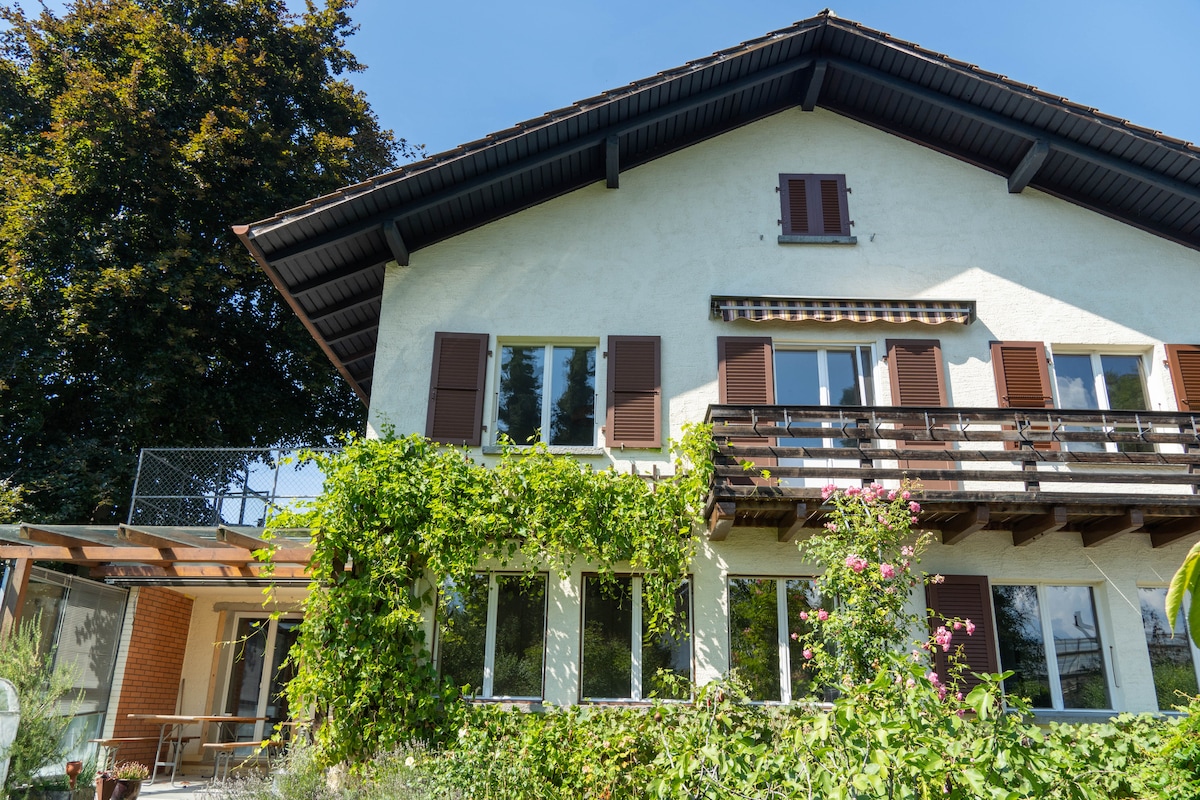 The exterior of the two-story home showcases a charming facade adorned with climbing greenery and blooming flowers. A balcony with shutters provides additional character, while large windows invite natural light. A spacious patio area is visible, enhancing the inviting atmosphere of the property.