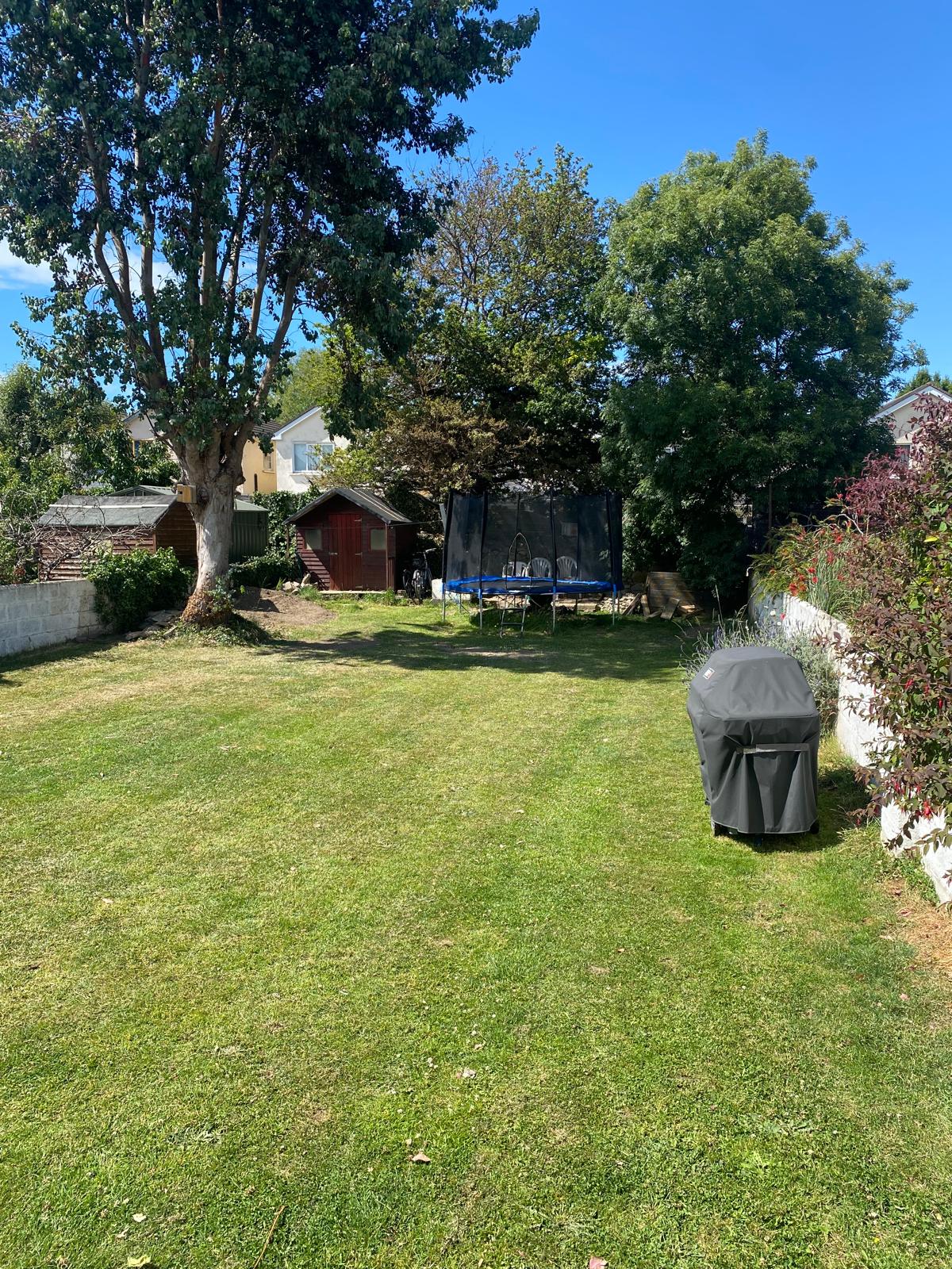A large rear garden features well-maintained grass and is oriented to receive abundant sunlight. A trampoline is positioned in the center, surrounded by trees, while a barbecue grill is set on one side, providing a space for outdoor activities.