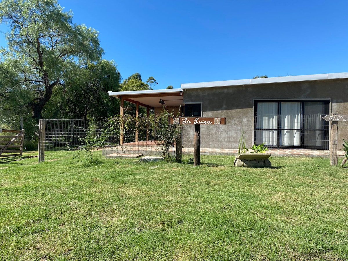 A single-story house is set against a clear blue sky, featuring a welcoming entry with a sign reading 'La Luisa.' Green grass surrounds the property, complemented by trees in the background. A spacious patio is visible, offering a relaxing outdoor space.