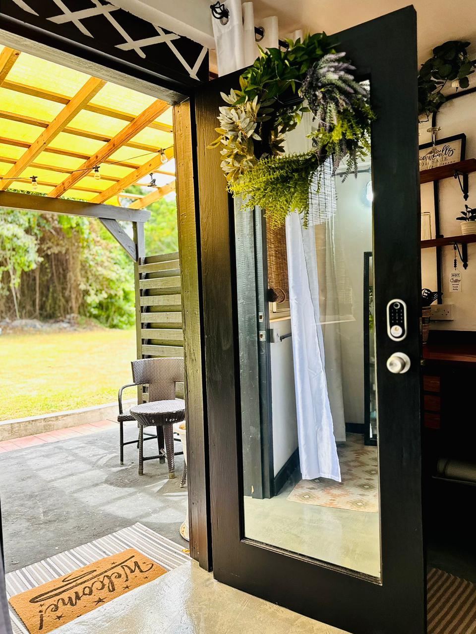 A glass door opens to a private patio, framed by greenery. A welcome mat sits at the entrance, and a small table with two chairs is visible on the patio. Natural light brightens the entrance, enhancing the connection with the outdoors.