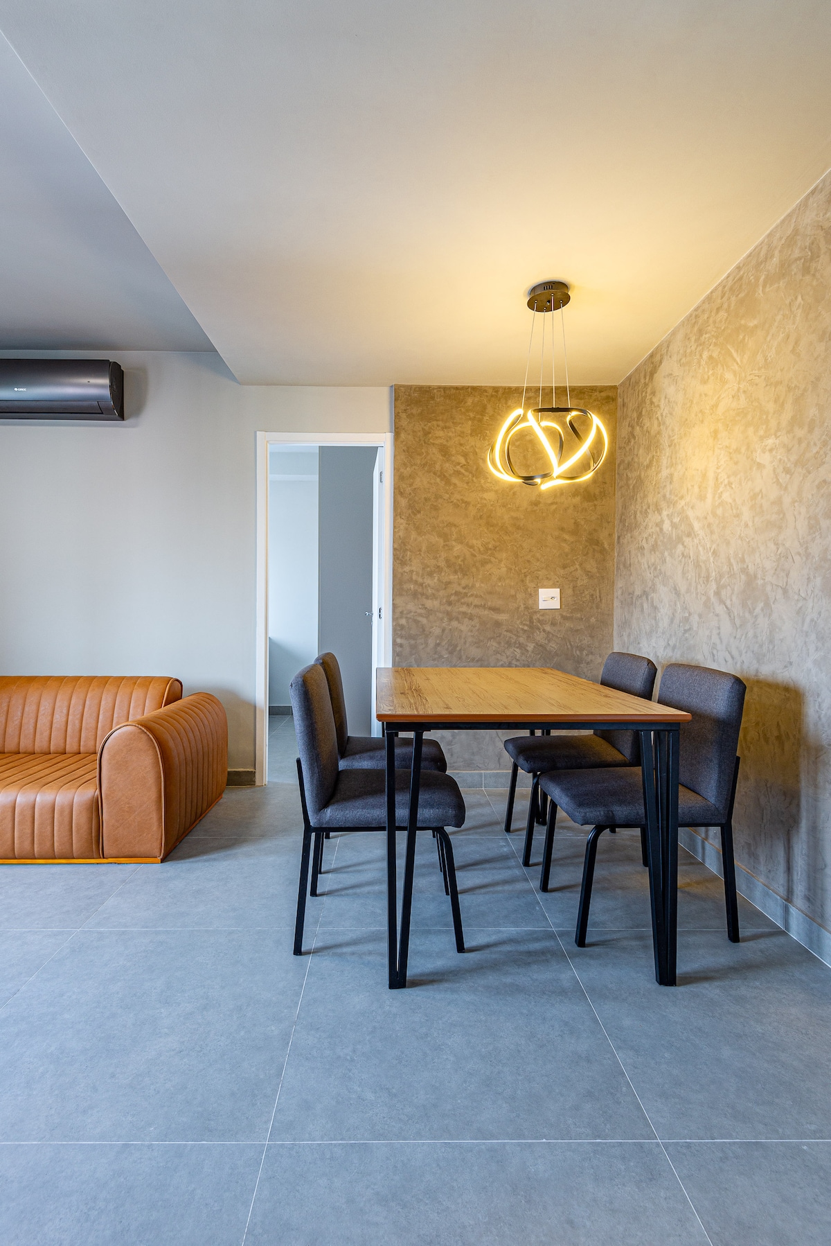 A dining area is featured with a wooden table surrounded by four black chairs. Above, a modern light fixture adds illumination. A cozy brown couch is visible in the background, complemented by a neutral-toned wall and a door leading to another room.
