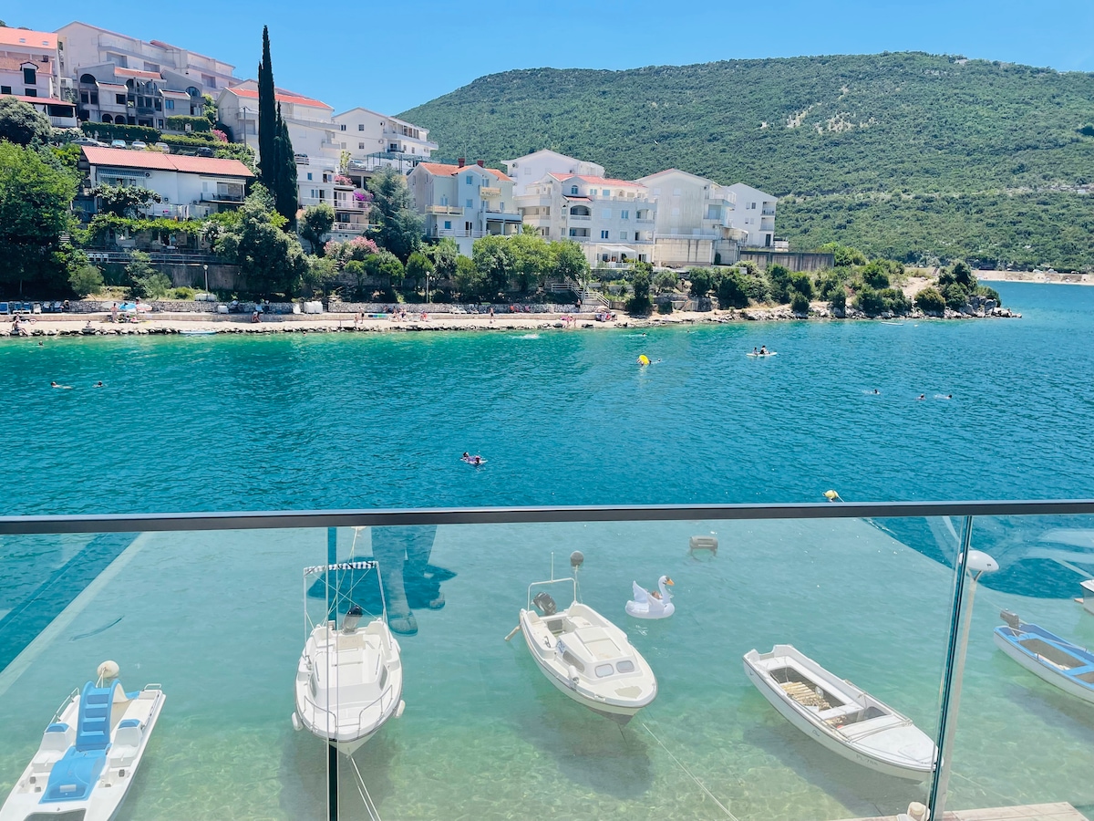 A waterfront view is showcased, featuring several boats bobbing gently in the clear water. The distant shoreline is lined with residences and greenery, while beachgoers can be seen enjoying the water. A glass balcony railing offers an unobstructed perspective of the vibrant landscape.