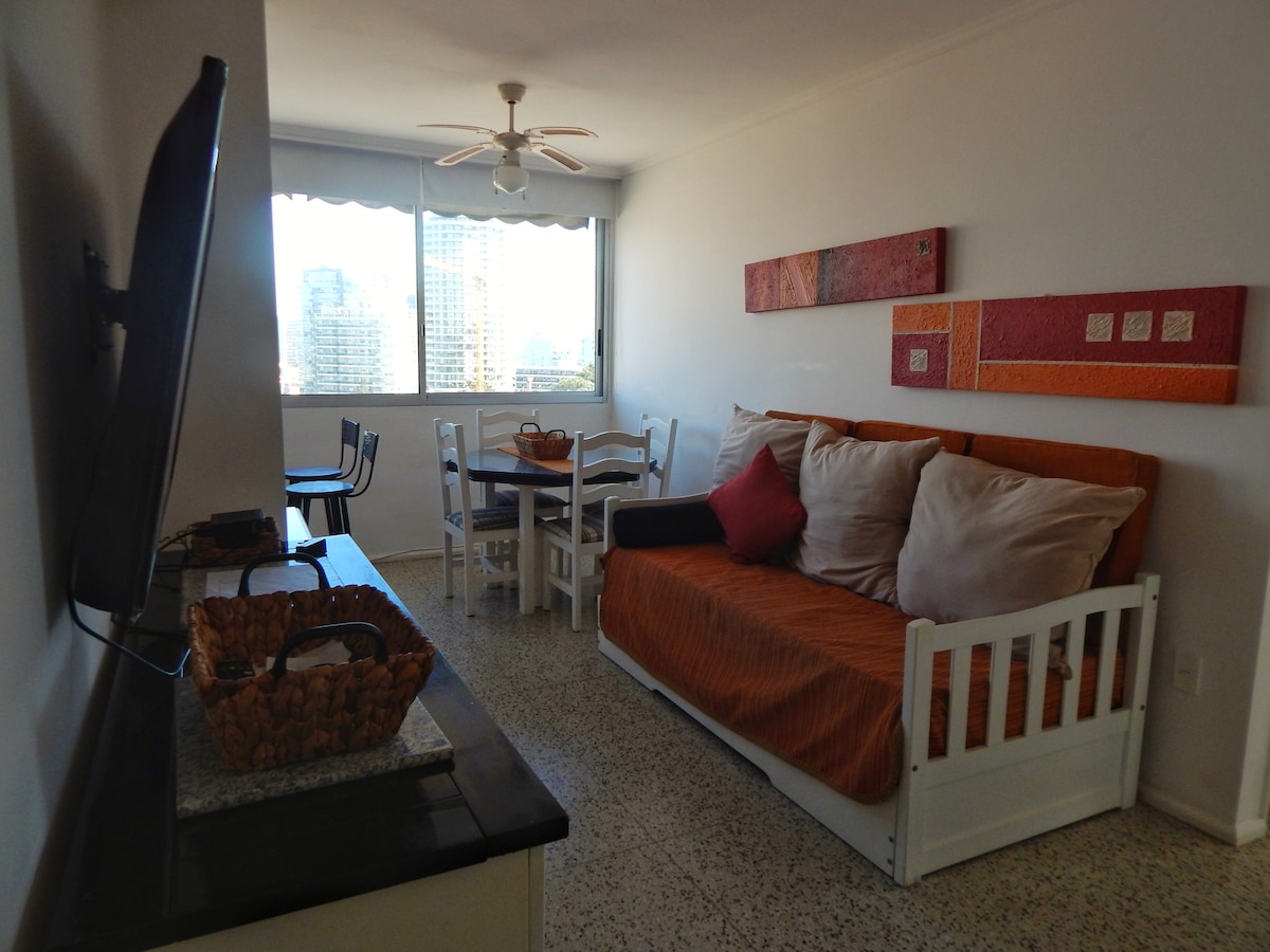 A living area is presented with a white futon adorned in orange and cream cushions, accompanied by a small dining table for four. A flat-screen TV is mounted on the wall, with natural light streaming through a large window that overlooks the cityscape.