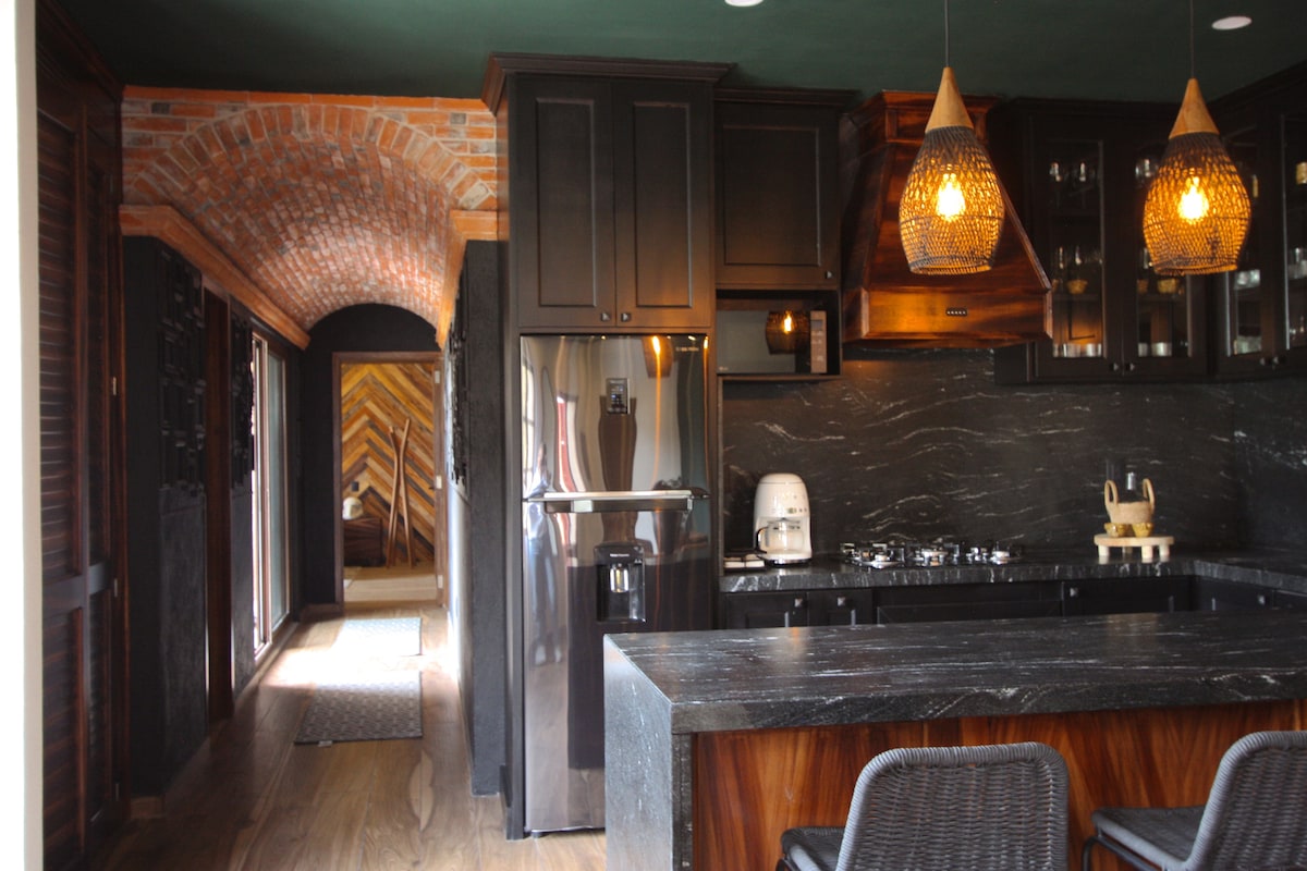 A modern kitchen is presented with dark cabinetry and a sleek black countertop. Two pendant lights hang above, providing illumination. Stainless steel appliances, including a refrigerator, are visible. The kitchen opens to a hallway with artistic wooden details, enhancing the overall design.