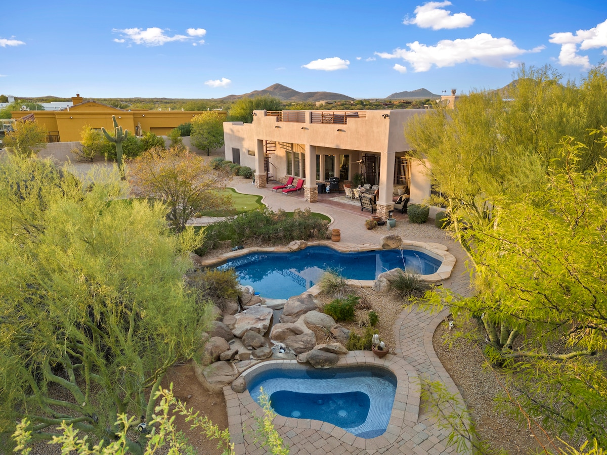 An aerial view of the villa showcases a spacious backyard featuring a natural rock spa and pool surrounded by lush desert landscaping. Comfortable seating areas are visible on the patio, and the mountain backdrop adds to the serene environment.