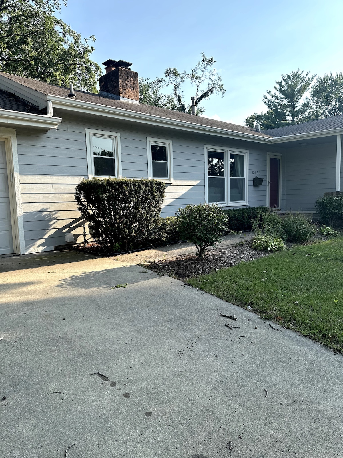 The exterior of a light gray house is presented, featuring neatly trimmed shrubs and a small garden area. Large windows allow for ample natural light. A paved driveway leads to the entrance, with a welcoming front porch visible. A wooded area provides a green backdrop.