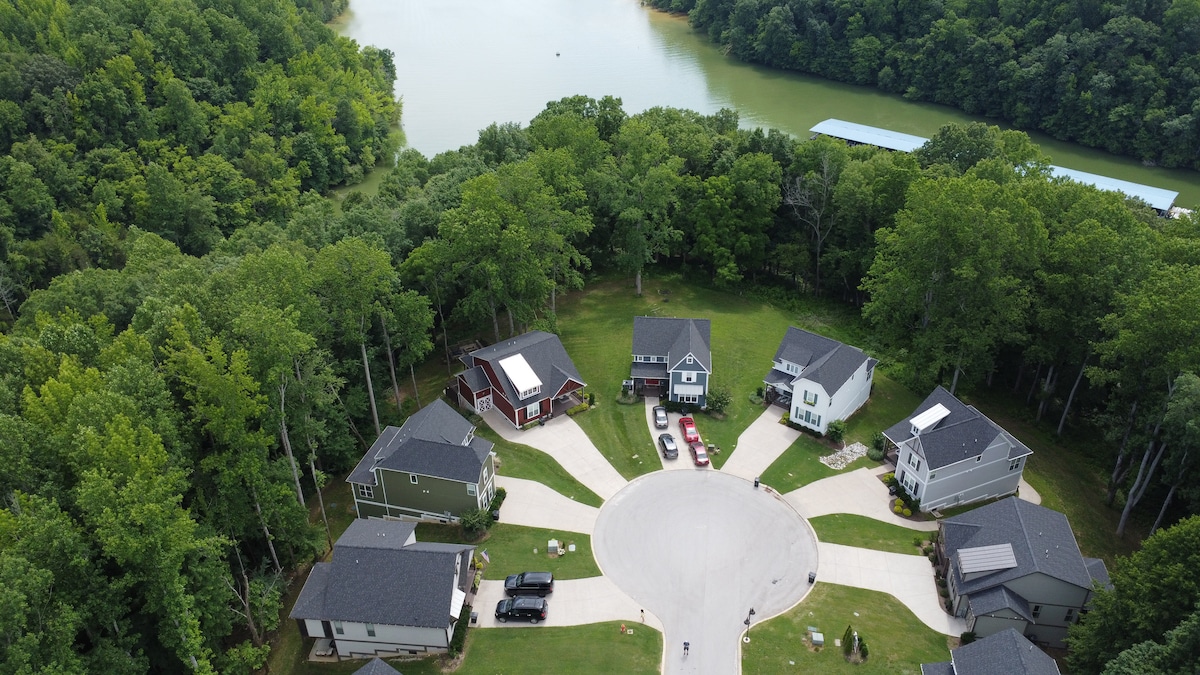 An aerial view captures a circular cul-de-sac surrounded by lush greenery and trees, with several homes visible. A serene lake glimmers in the background, enhancing the peaceful neighborhood setting.
