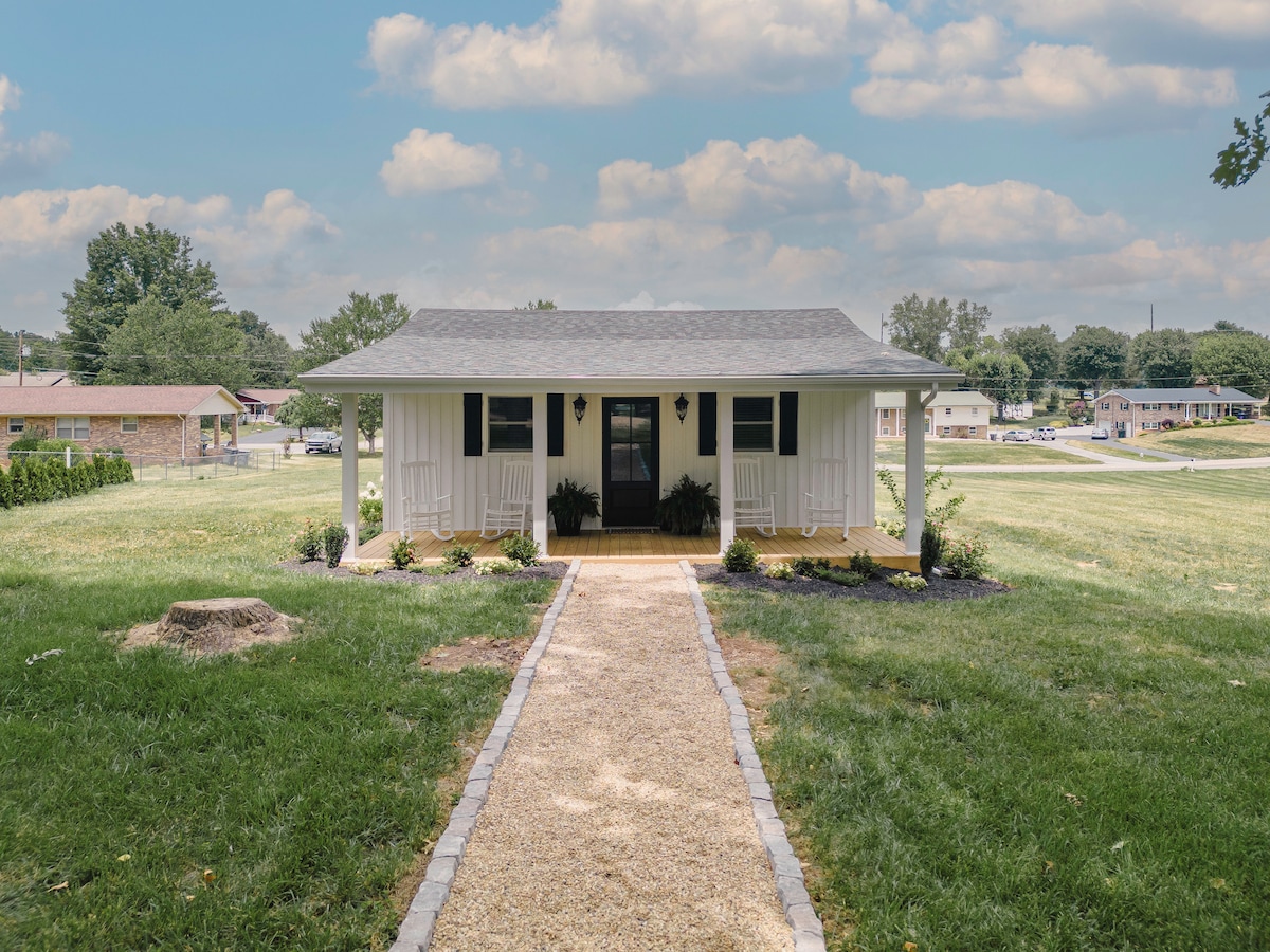 The entrance of the cottage is framed by a well-maintained pathway lined with gravel. Flanking the front door, two inviting rocking chairs sit on the porch, providing a view of the surrounding landscape and greenery.