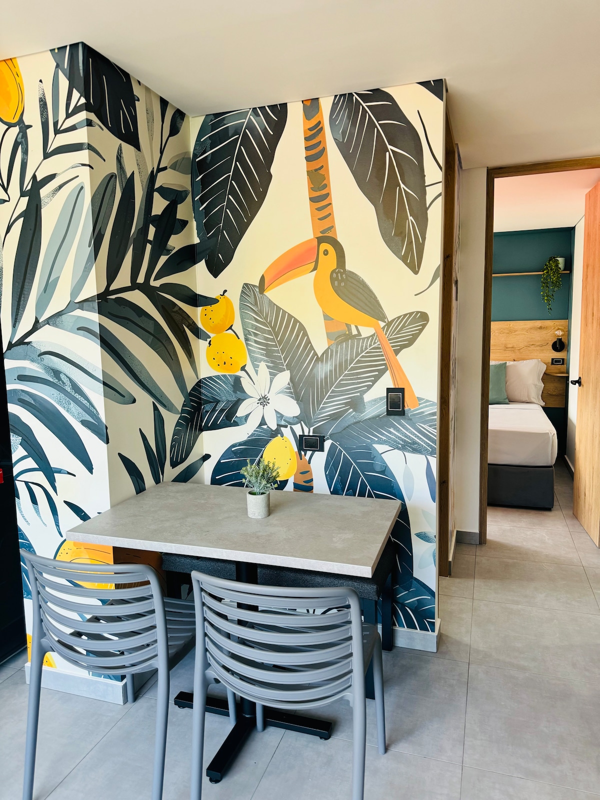 A vibrant dining area is showcased with a table and two gray chairs, set against a striking mural featuring tropical foliage and a toucan. The decor highlights lively colors, and the doorway in the background leads into the bedroom area.