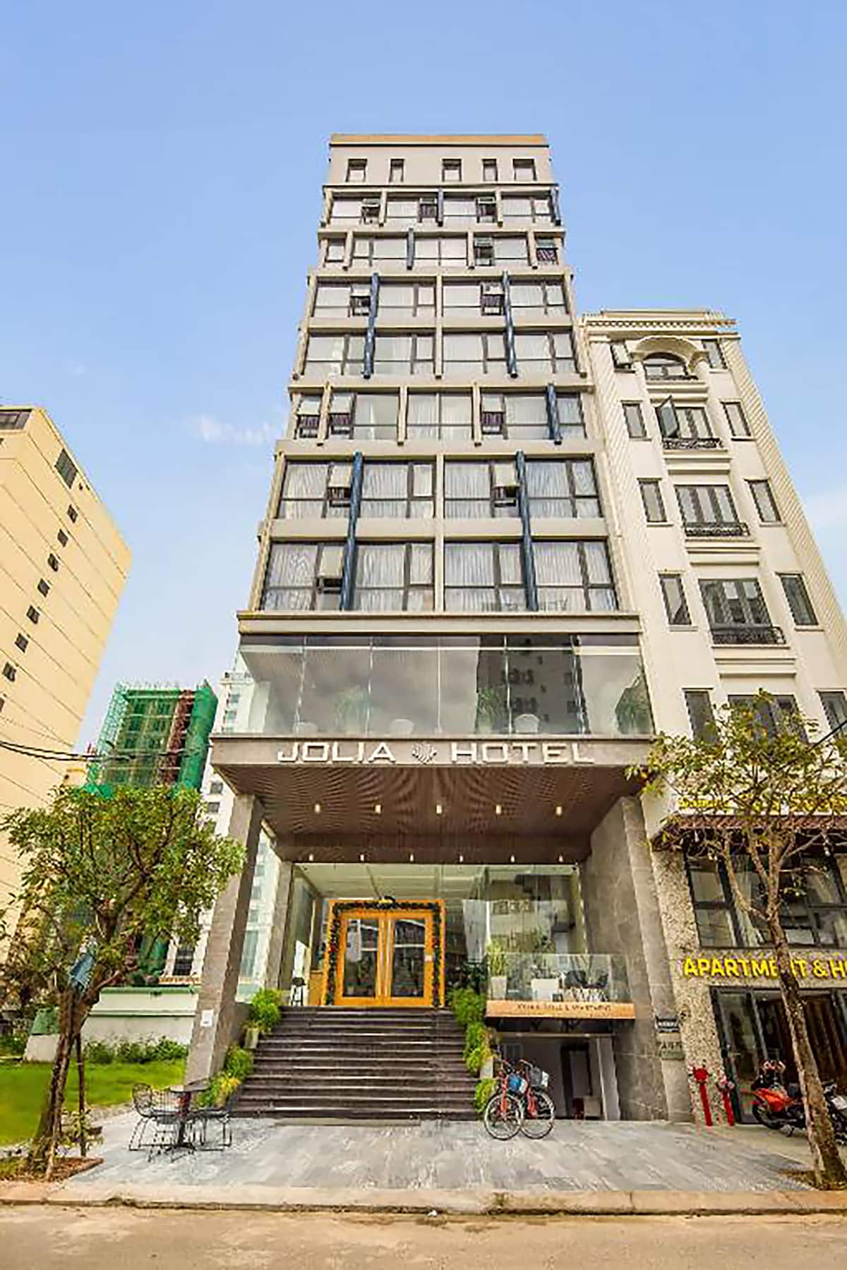 The Jolia Hotel Danang Beach is showcased with a modern facade featuring large glass windows and a welcoming entrance. The multi-story building is flanked by neatly trimmed trees on the front, highlighting the hotel’s accessible and inviting atmosphere.