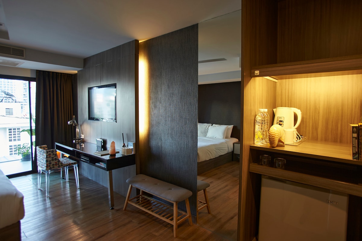 An open living space is displayed with a dark wood accent wall and a cozy sleeping area beyond. A flat-screen TV is mounted on the wall opposite a sleek desk and chair. A small kitchenette area is visible with a kettle and a refrigerator.