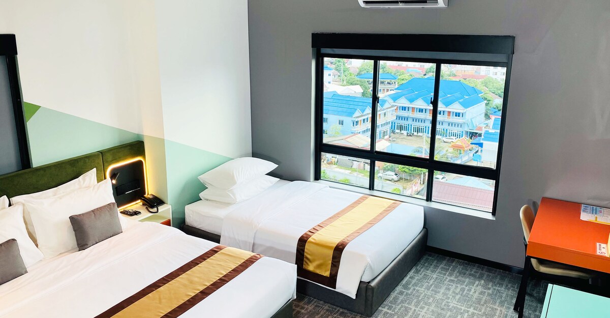 The family room features a large king-size bed alongside a single bed, each adorned with neutral linens. A spacious window provides a view of the cityscape, while a small desk is positioned against the wall, offering a functional work area.