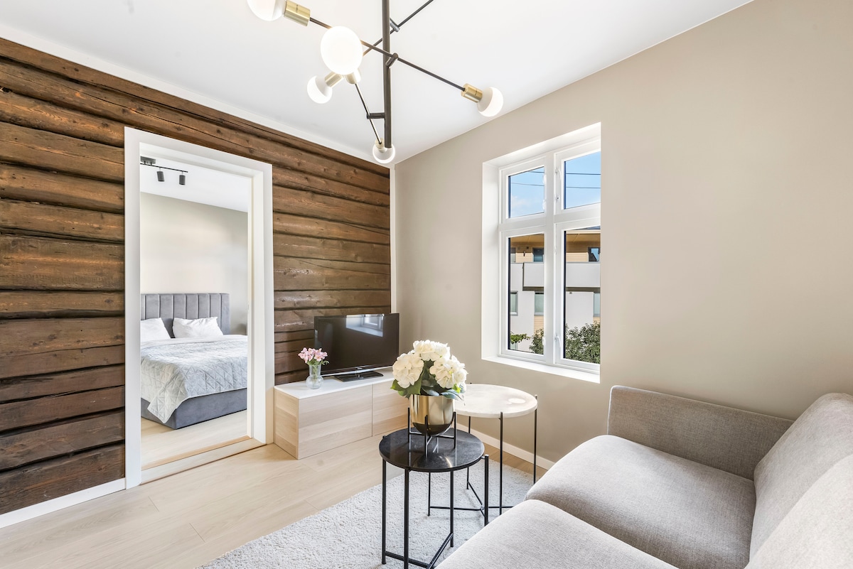 A modern living space features a stylish light fixture and a cozy sofa, set against a backdrop of wooden wall panels. Natural light filters through large windows, illuminating a small coffee table and a television. A doorway connects to a bedroom area, visible in the background.