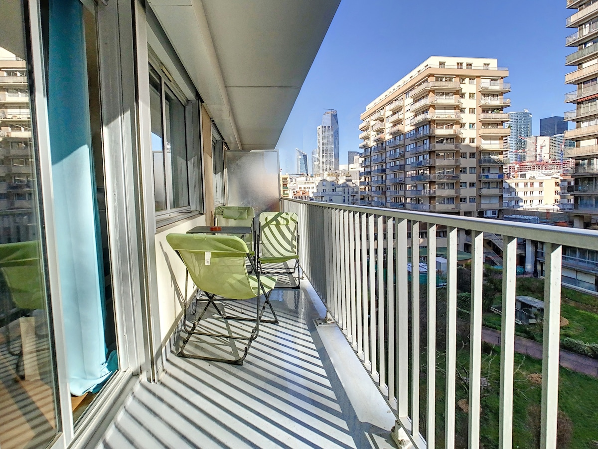 A balcony features two green chairs arranged for outdoor seating, with a railing providing safety. Natural light creates a pleasing ambiance, while city buildings in the background highlight the urban setting. The view includes green spaces below, contributing to the apartment's calm environment.